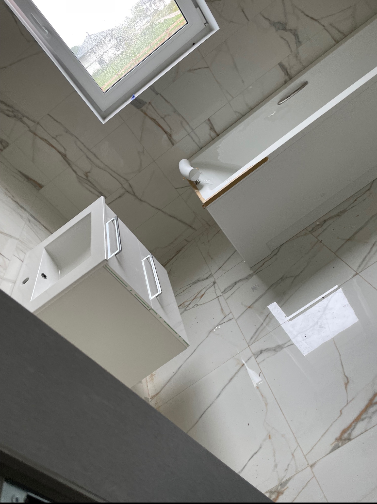 Nowoczesna łazienka z białą umywalką, wanną i marmurową podłogą, widziana z perspektywy sufitowego okna. Minimalistyczny design, jasne wnętrze.