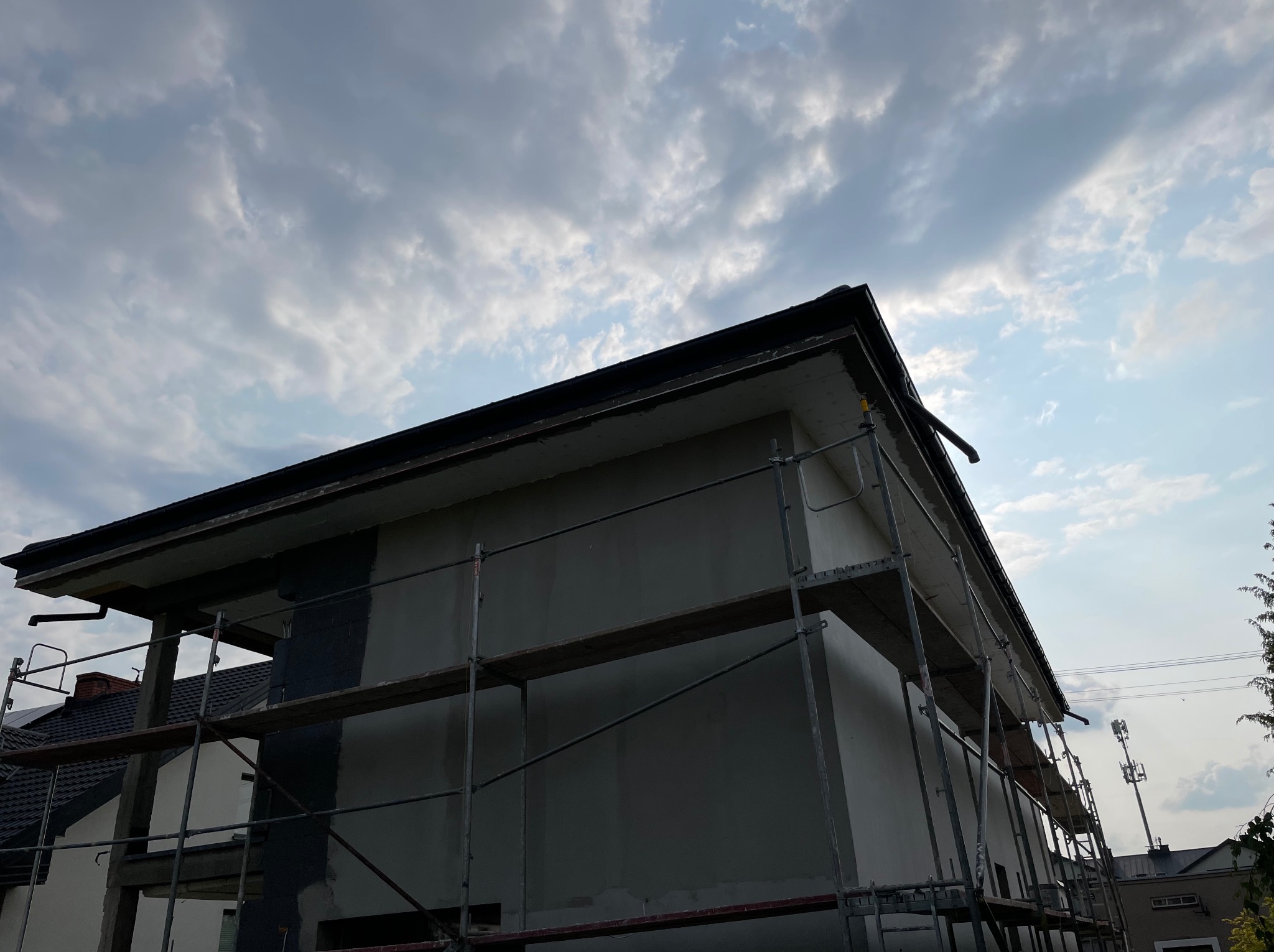 Dom w trakcie tynkowania elewacji, widoczne rusztowania. Częściowo otynkowana ściana budynku na tle pochmurnego nieba. Detal: rynna przy dachu.