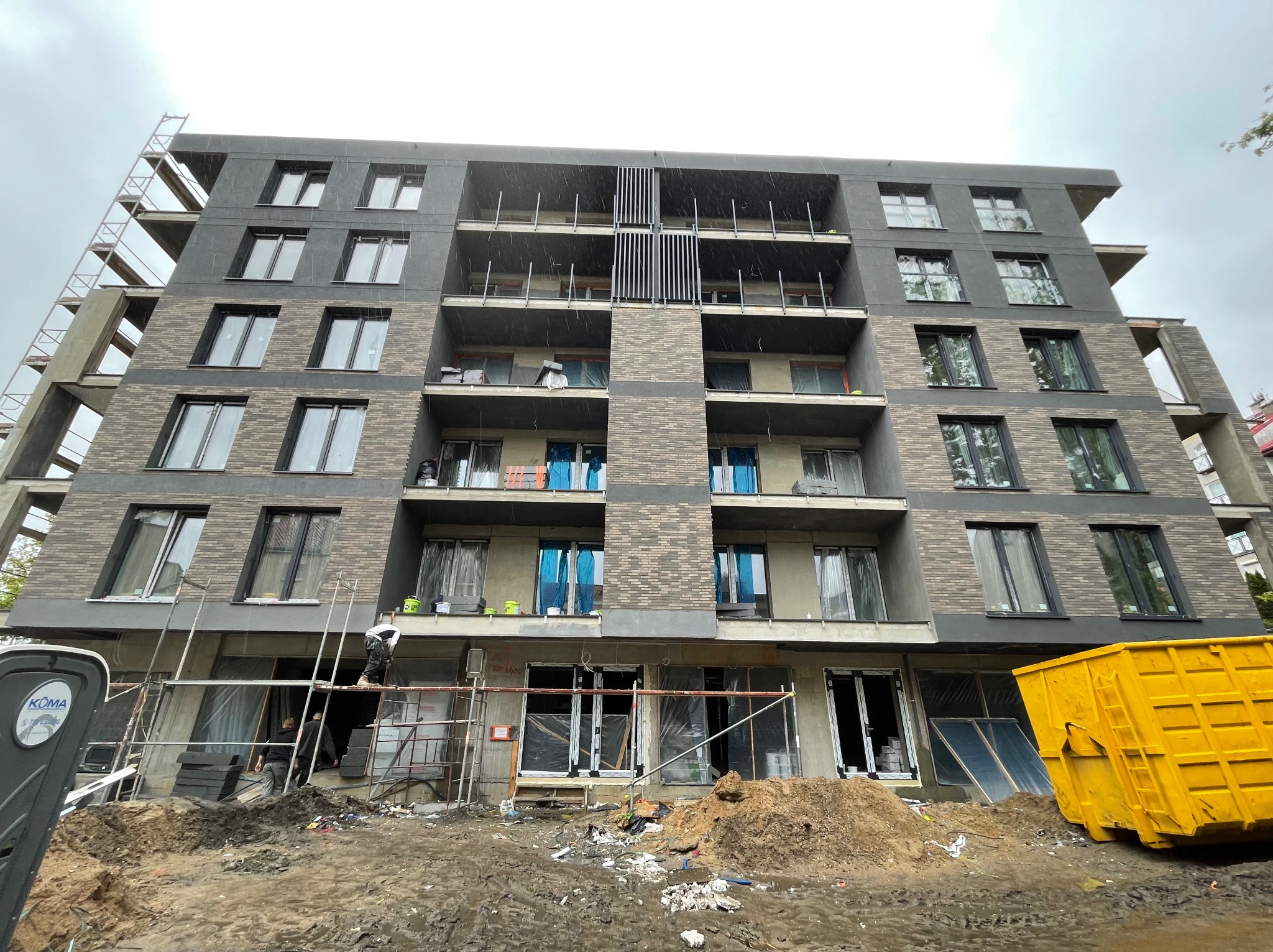 Wielopiętrowy budynek z elewacją w trakcie realizacji. Widoczne rusztowania, pracownicy i kontener na odpady. Część okien zabezpieczona folią.