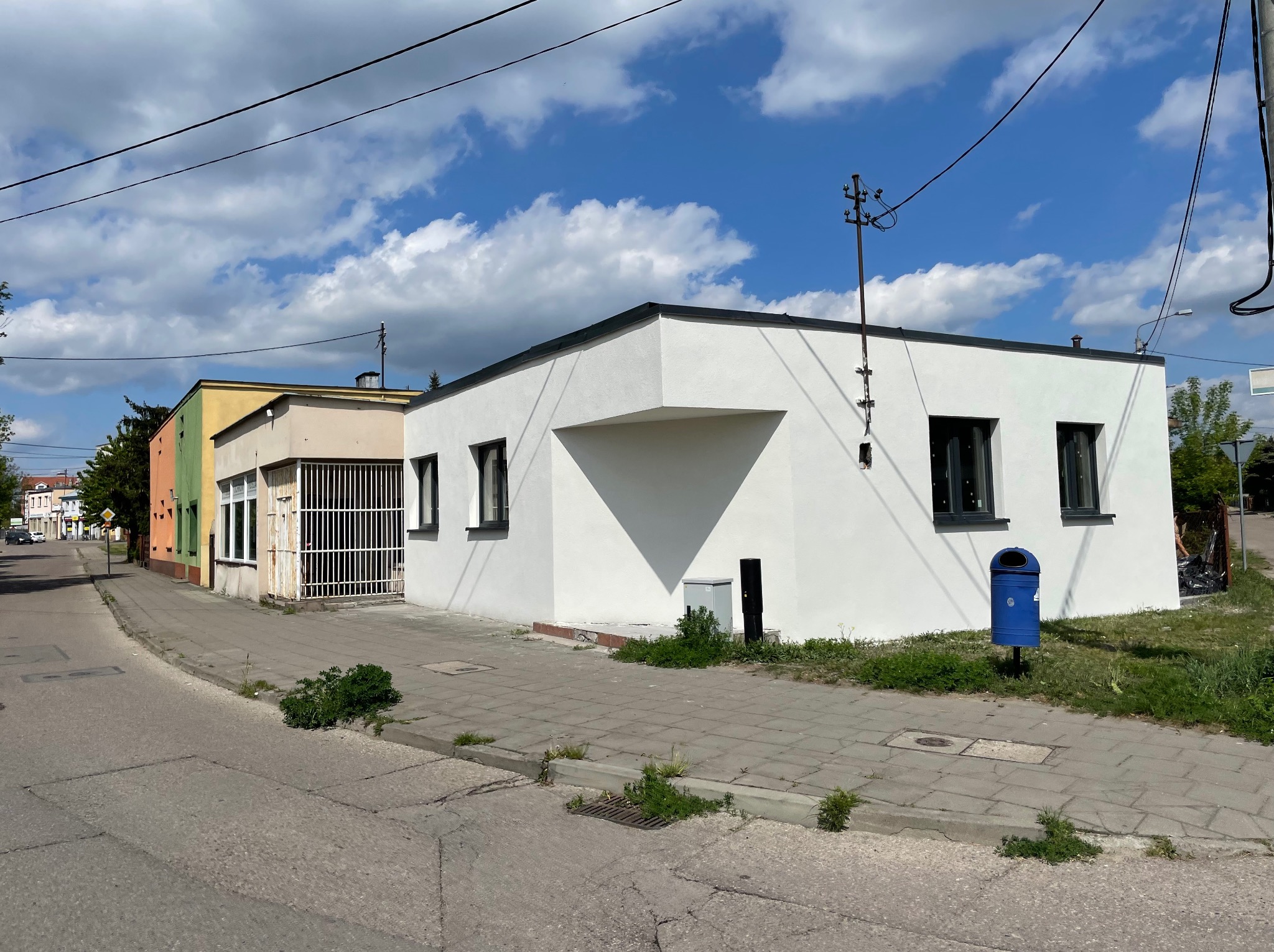 Biały, odnowiony budynek z ciemnymi oknami i niebieskim koszem na śmieci na chodniku, kontrastujący z błękitnym niebem i chmurami.