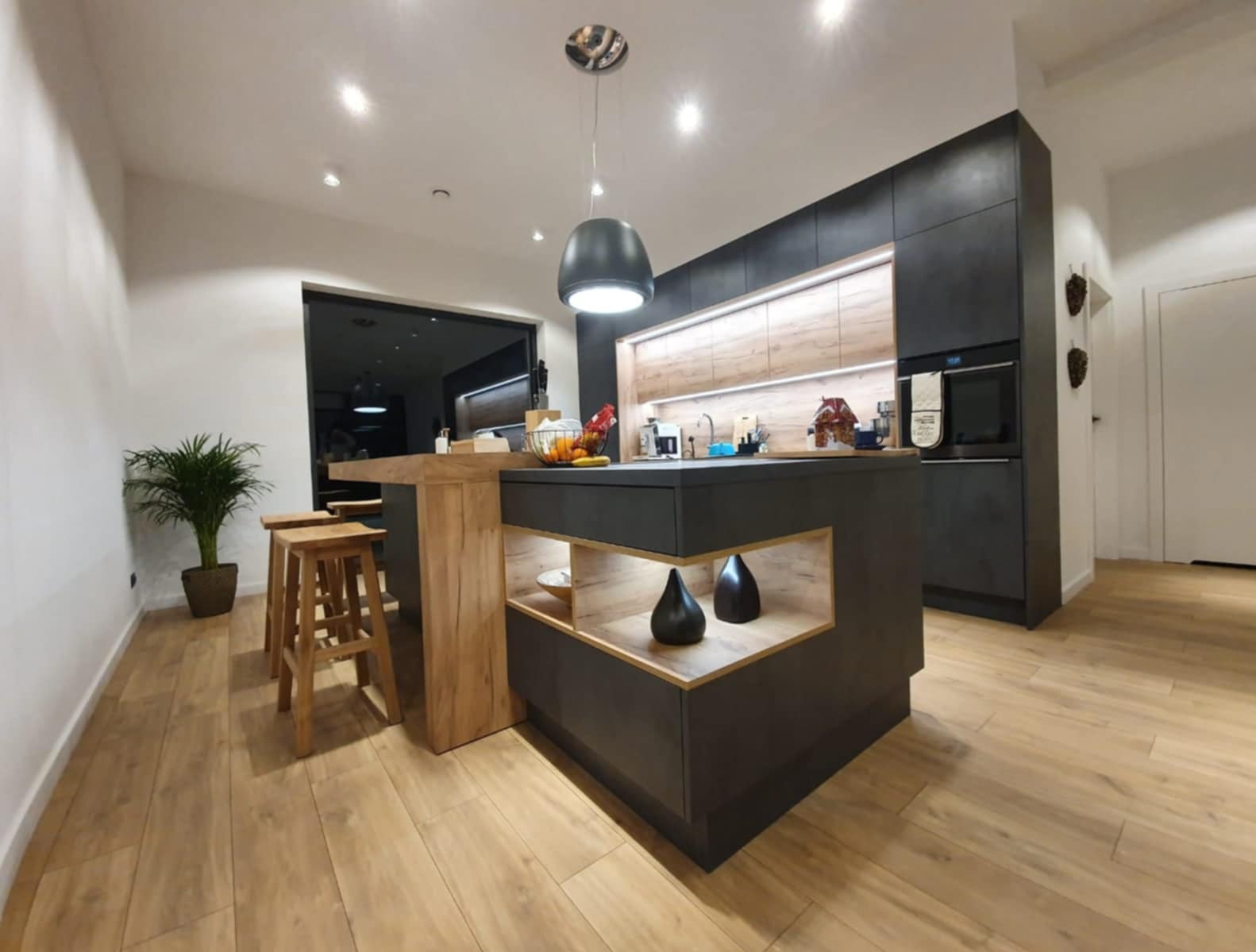 Nowoczesna kuchnia z wyspą i drewnianymi hokerami. Ciemnoszare fronty szafek kontrastują z drewnianymi elementami. Oświetlenie LED podkreśla charakter wnętrza.