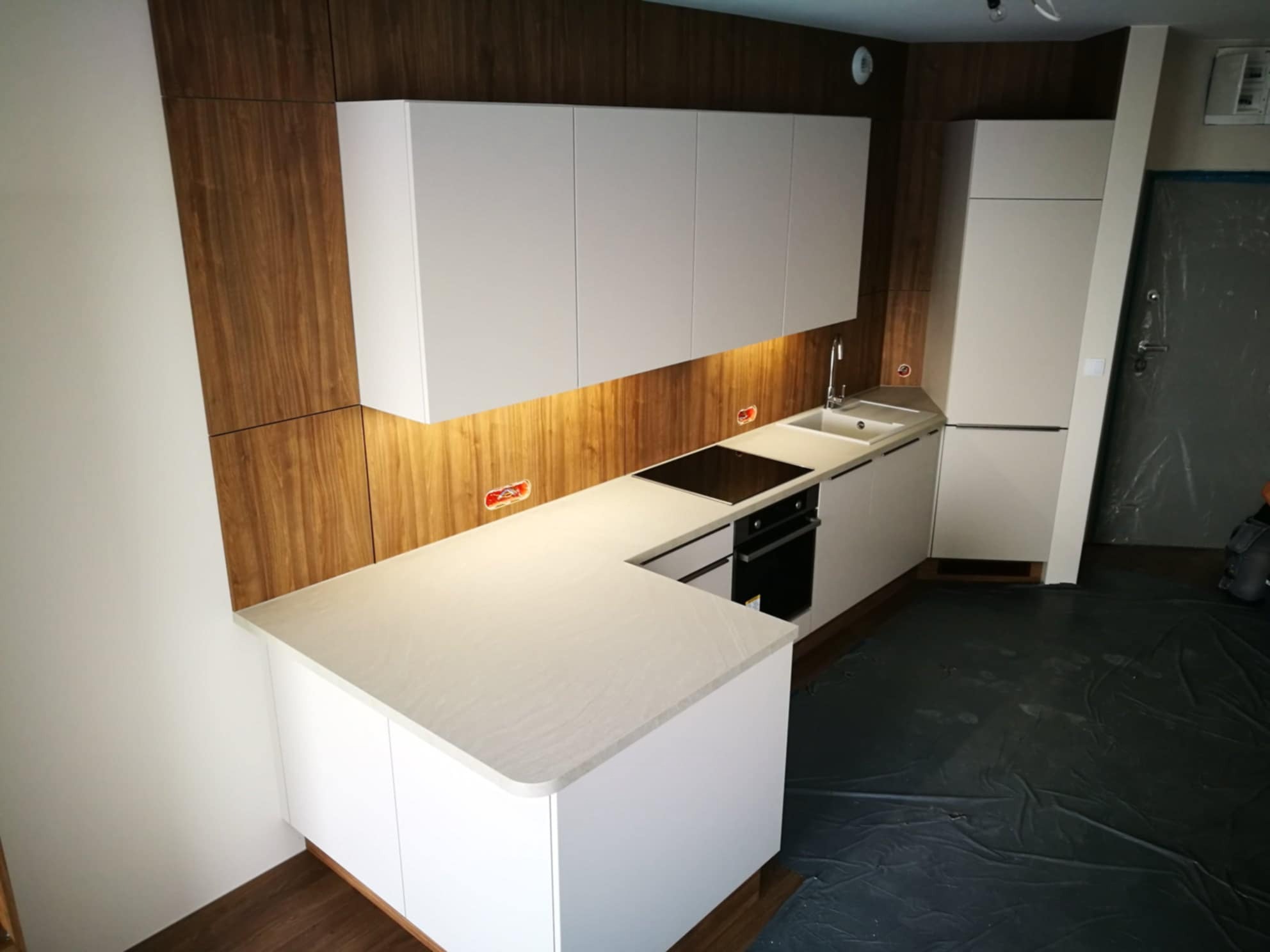 Nowoczesna kuchnia z białymi szafkami i drewnianym panelem ściennym, oświetlona listwą LED pod szafkami. Uwagę zwraca czarny blat kuchenki indukcyjnej.