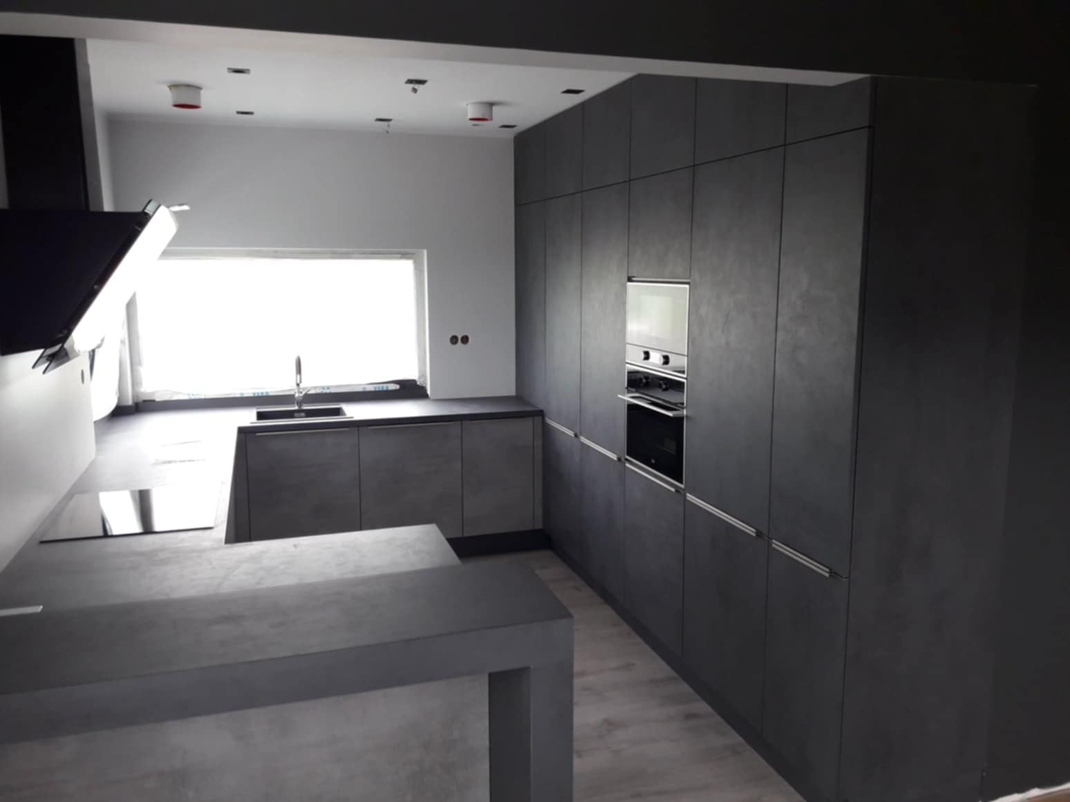 Nowoczesna kuchnia w odcieniach szarości z wyspą, oświetleniem punktowym i wbudowanym piekarnikiem, minimalistyczny design.