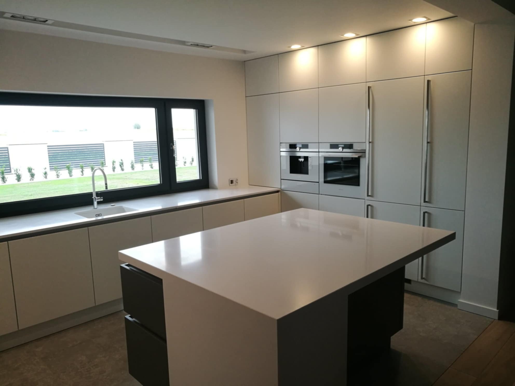 Nowoczesna kuchnia z wyspą na tle okna. Jasne, gładkie fronty szafek i zabudowane sprzęty AGD tworzą minimalistyczny design. Uwagę przykuwa duża powierzchnia blatu.