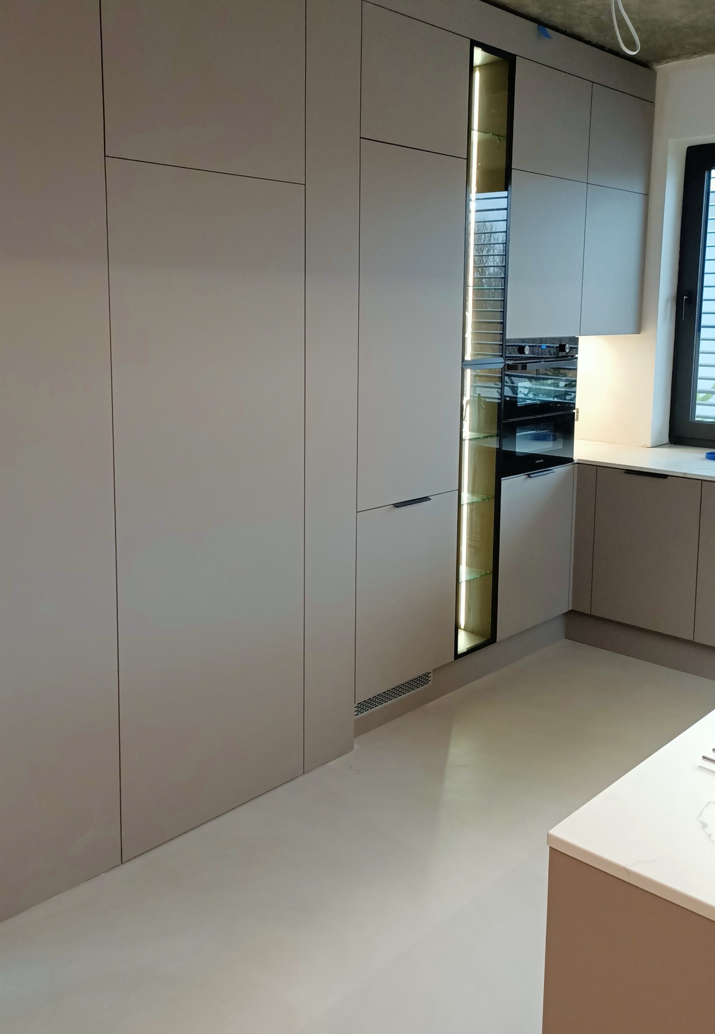 Jasnoszara zabudowa kuchenna z oświetleniem LED w witrynie i piekarnikiem wkomponowanym w szafkę. Minimalistyczny design, nowoczesne wykończenie.
