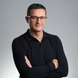 Portret mężczyzny w okularach i czarnej koszulce polo z logo BOSS, z założonymi rękami na tle szarego studia. Uśmiechnięty, pewny siebie wyraz twarzy.