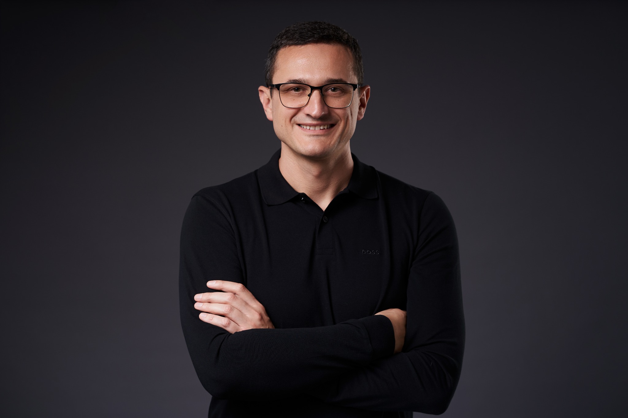 Uśmiechnięty mężczyzna w okularach i czarnej koszulce polo z długim rękawem, pozujący na szarym tle z założonymi rękami.