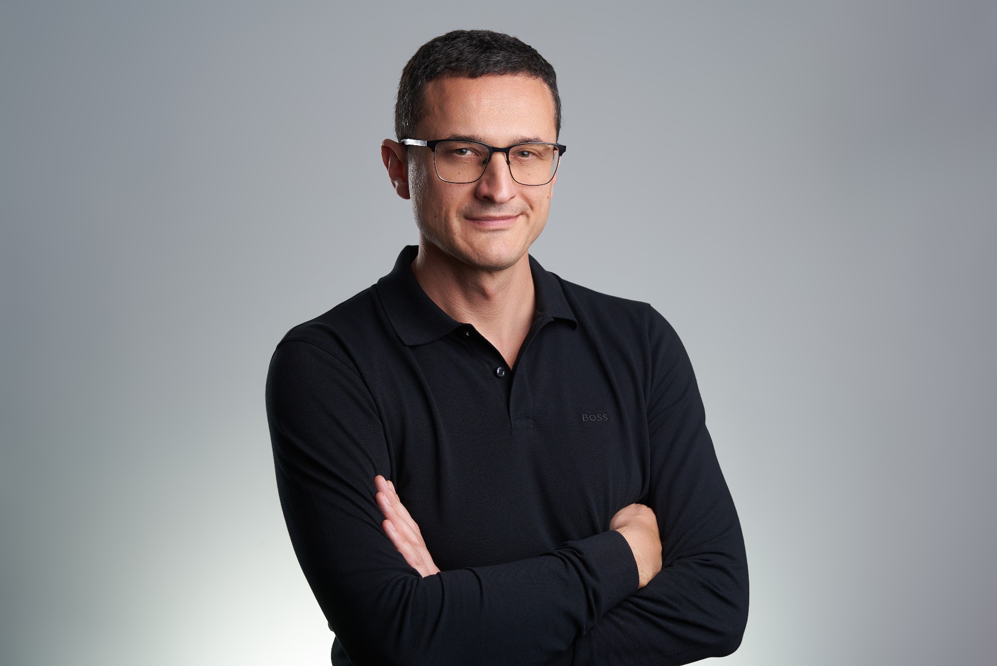 Portret mężczyzny w okularach i czarnej koszulce polo z logo BOSS, z założonymi rękami na tle szarego studia. Uśmiechnięty, pewny siebie wyraz twarzy.