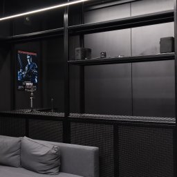KBD Kacper Błaszczyk Design - Czarna, metalowa konstrukcja regału z ażurowym blatem, w tle plakat filmowy i sofa. Minimalistyczne wnętrze w ciemnej tonacji, oświetlone listwą LED.
