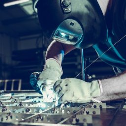 KBD Kacper Błaszczyk Design - Spawanie metalowej konstrukcji kratowej w warsztacie. Spawacz w masce ochronnej i rękawicach łączy elementy, iskrzy. W tle zarys wyposażenia warsztatowego.