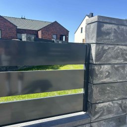 KBD Kacper Błaszczyk Design - Nowoczesne ogrodzenie z poziomych, szarych paneli metalowych i kamiennych słupków na tle domów i zielonego trawnika, w słoneczny dzień.