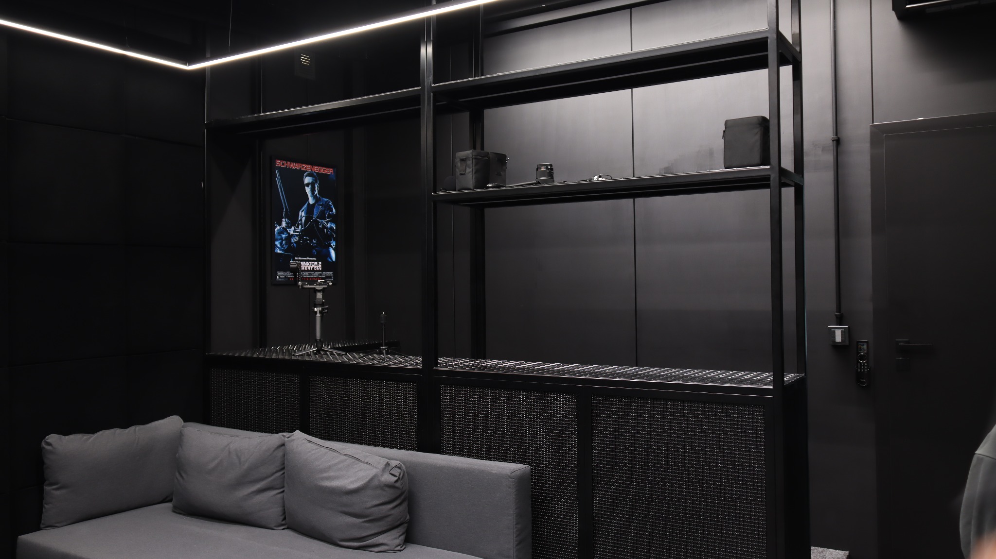 Czarna, metalowa konstrukcja regału z ażurowym blatem, w tle plakat filmowy i sofa. Minimalistyczne wnętrze w ciemnej tonacji, oświetlone listwą LED.