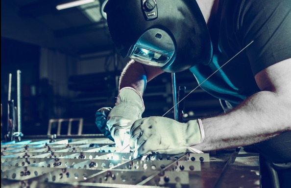 Spawanie metalowej konstrukcji kratowej w warsztacie. Spawacz w masce ochronnej i rękawicach łączy elementy, iskrzy. W tle zarys wyposażenia warsztatowego.