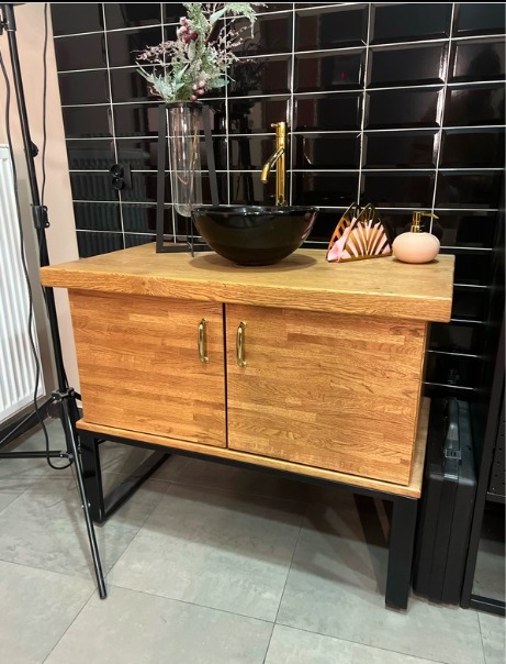 Szafka łazienkowa z dębowego drewna z czarną umywalką i złotą armaturą na tle czarnych płytek. Uwagę przykuwają złote uchwyty oraz nowoczesny design.