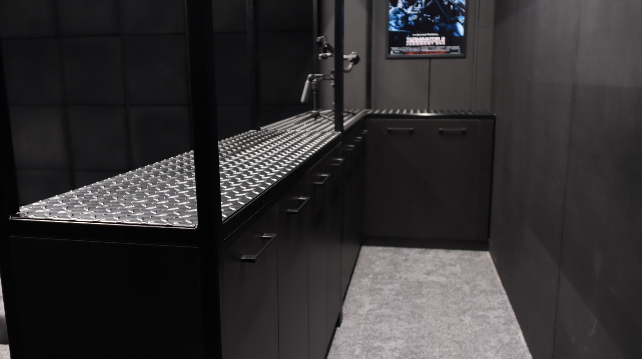 Metalowa zabudowa kuchenna w ciemnej tonacji z blatem z blachy ryflowanej, widoczne szafki z uchwytami, na ścianie plakat filmowy. Industrialny styl.