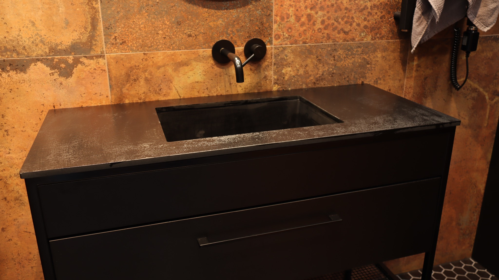 Czarna szafka łazienkowa z umywalką w stylu industrialnym, na tle ściany w odcieniach rdzy. Minimalistyczna bateria ścienna i heksagonalna podłoga.