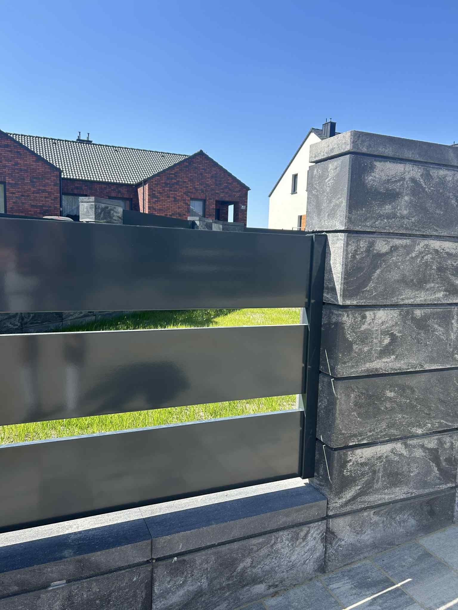 Nowoczesne ogrodzenie z poziomych, szarych paneli metalowych i kamiennych słupków na tle domów i zielonego trawnika, w słoneczny dzień.