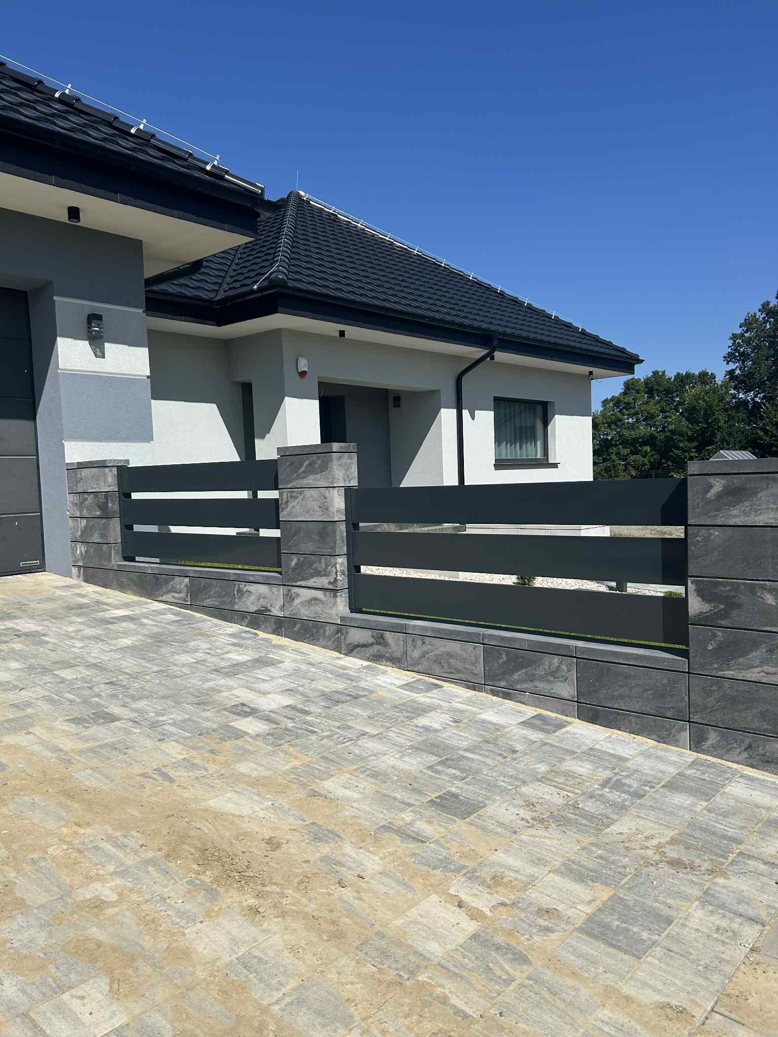 Nowoczesne ogrodzenie posesji z poziomych, metalowych paneli i kamiennych słupków w Warszawie, widoczne z poziomu brukowanego podjazdu.