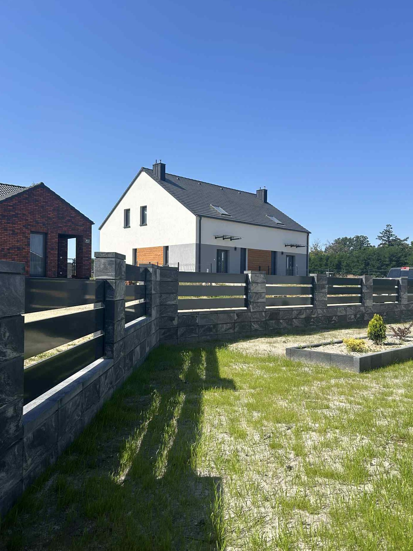 Nowoczesne ogrodzenie panelowe z szarego kamienia łupanego, z poziomymi, metalowymi przęsłami, na tle domu jednorodzinnego i błękitnego nieba.