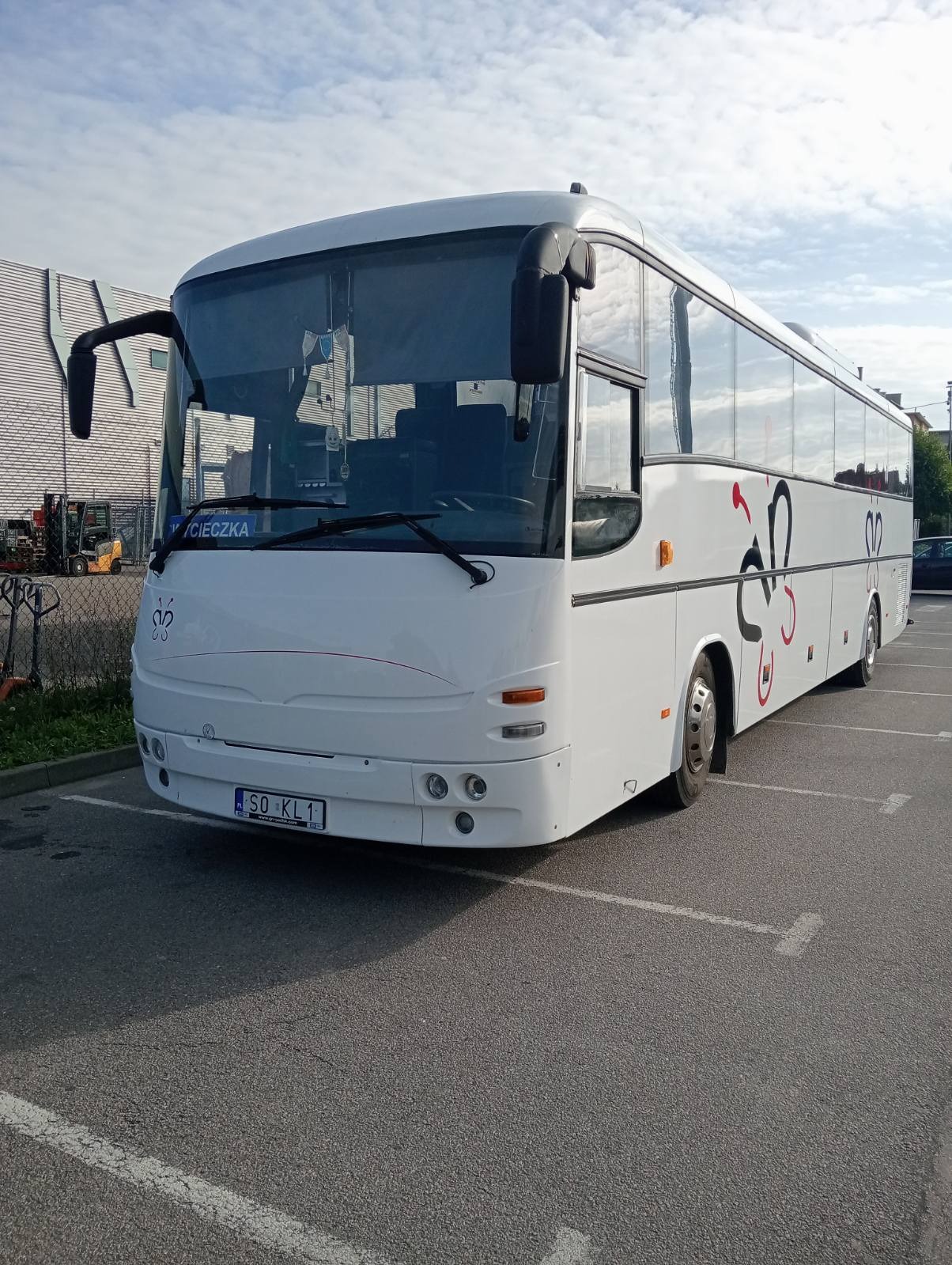 Biały autokar z dekoracyjnym motywem parkuje na parkingu. Widoczny przód pojazdu z tablicą rejestracyjną i oznaczeniem 'Cieczka' nad przednią szybą.