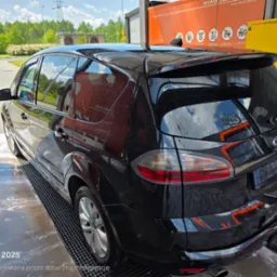Czarny van Ford S-Max po myciu w myjni automatycznej, widok z tyłu, mokry lakier odbija otoczenie. W tle zieleń drzew i błękitne niebo.