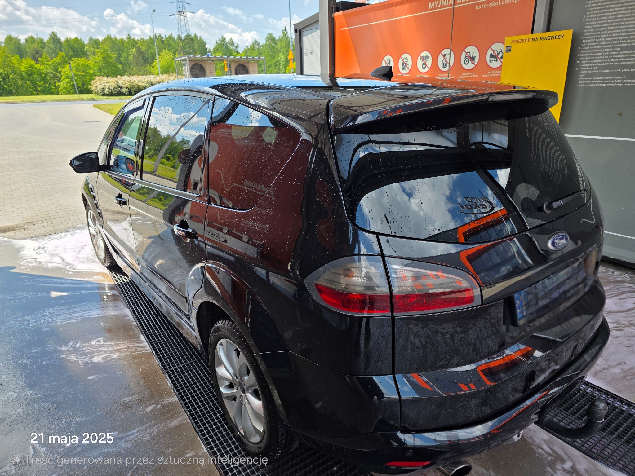 Czarny van Ford S-Max po myciu w myjni automatycznej, widok z tyłu, mokry lakier odbija otoczenie. W tle zieleń drzew i błękitne niebo.