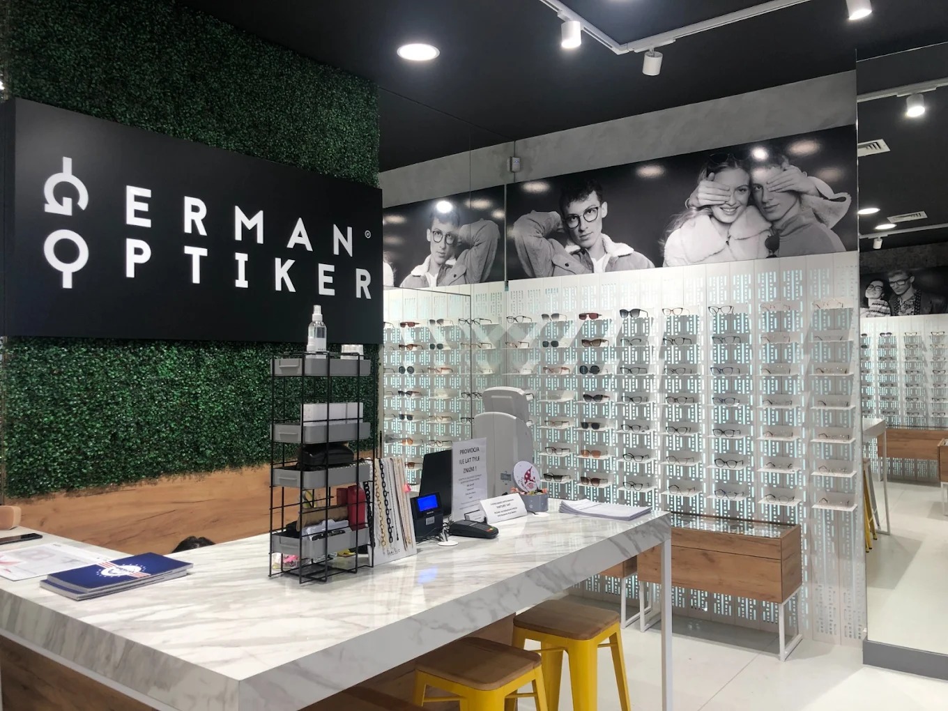 Wnętrze salonu optycznego z ekspozycją okularów na ścianie, marmurowym blatem recepcyjnym i dekoracją z zielonej roślinności. Nad blatem widoczny szyld z nazwą firmy.