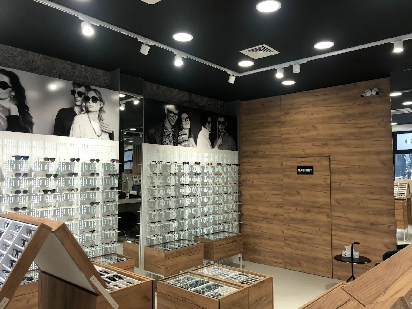 Salon optyczny z ekspozycją okularów na tle czarno-białych fotografii modeli. Drewniane regały i oświetlenie punktowe tworzą elegancki wystrój.