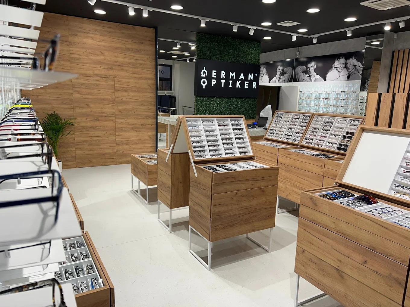 Salon optyczny 'Herman Optiker': drewniane ekspozytory z okularami, zielona ściana z logo, subtelne oświetlenie, elegancki wystrój.