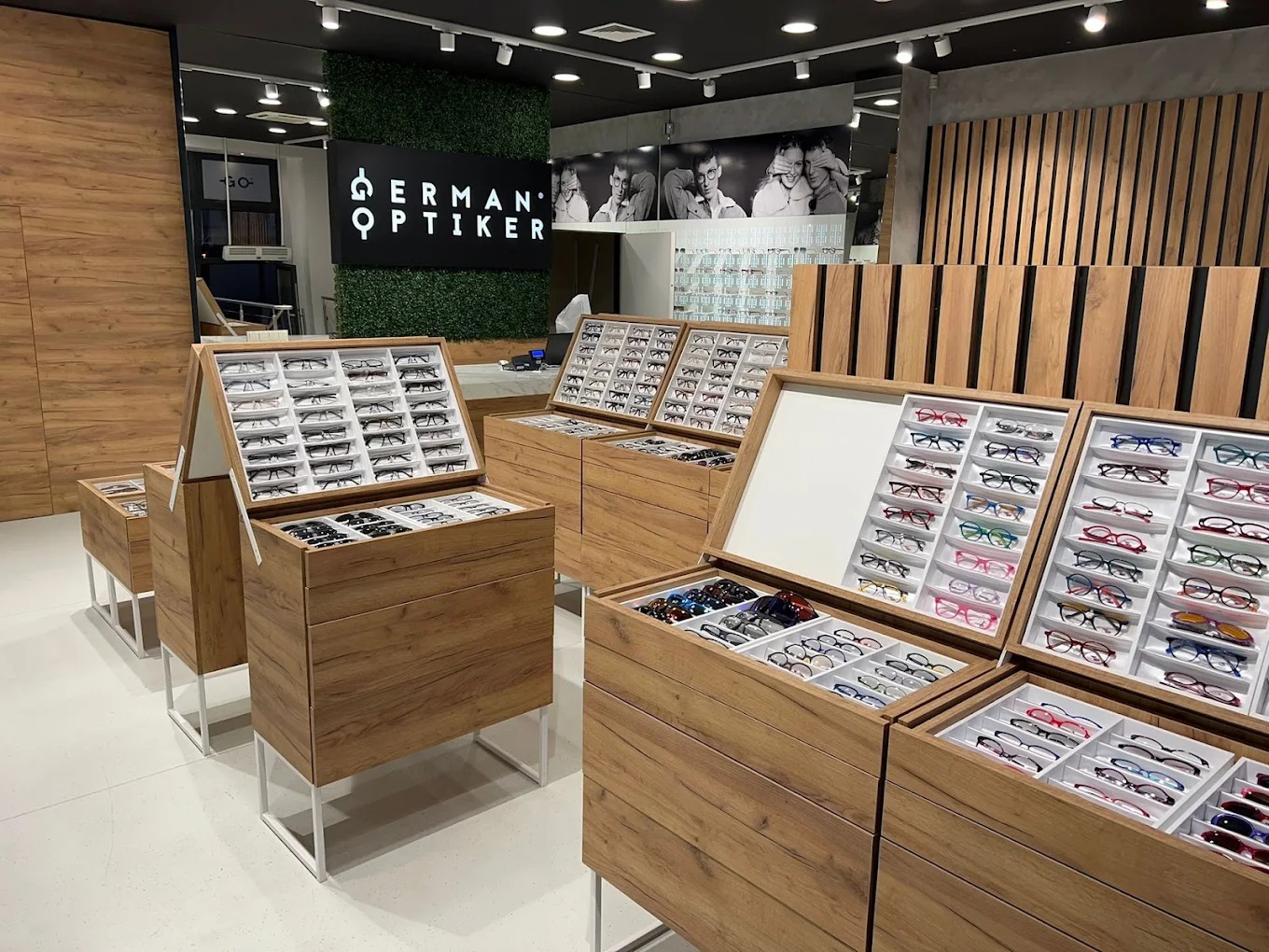 Salon optyczny z ekspozycją okularów korekcyjnych i przeciwsłonecznych w drewnianych gablotach, logo firmy na zielonej ścianie, czarno-białe zdjęcie na ścianie w tle.