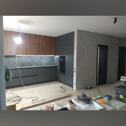 Fachowiec budowlany - Nowoczesna kuchnia w trakcie montażu: szare szafki dolne i drewniane górne, surowa ściana, kartony na podłodze. Widoczne narzędzia i akcesoria montażowe.