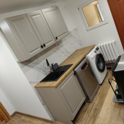 Fachowiec budowlany - Jasna kuchnia z drewnianym blatem, marmurową ścianą i sprzętami AGD. Uwagę przykuwają szare szafki z czarnymi uchwytami i drewnopodobne płytki na podłodze.