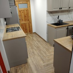 Fachowiec budowlany - Jasna kuchnia z drewnianą podłogą ułożoną w jodełkę, szare szafki z czarnymi uchwytami i drewniany blat. Widoczny fragment lodówki i drzwi z szybą.