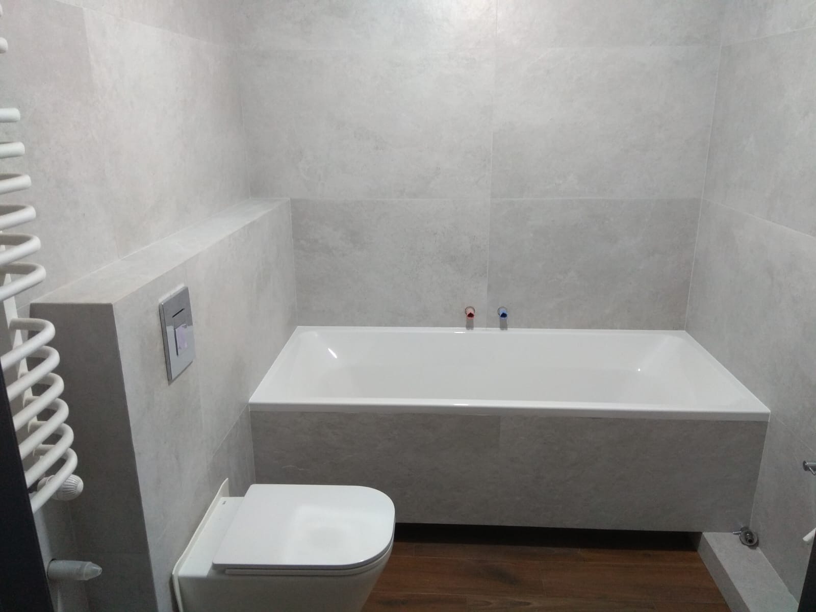 Jasna łazienka po remoncie: wanna obudowana płytkami, podwieszana toaleta, grzejnik drabinkowy. Minimalistyczny design, szare płytki, drewnopodobna podłoga.