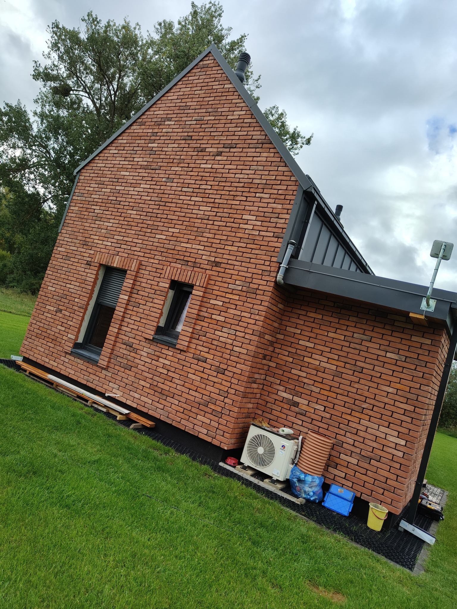 Ceglana elewacja domu z czarnymi oknami i dachem, widoczna z poziomu trawnika. Na dole klimatyzator i akcesoria budowlane. Ujęcie z lekkiego ukosa.