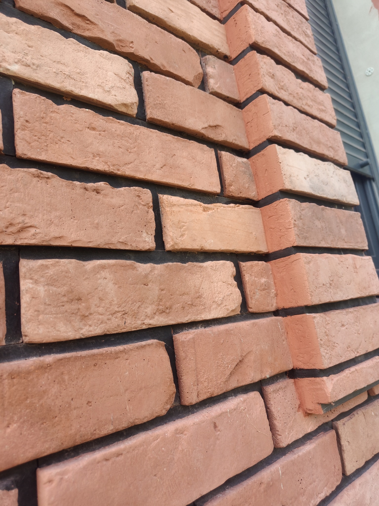 Ściana z klinkieru w odcieniach czerwieni i beżu, ułożona z przesunięciem, widoczna z bliska pod kątem, z ciemną fugą i fragmentem okna w tle.