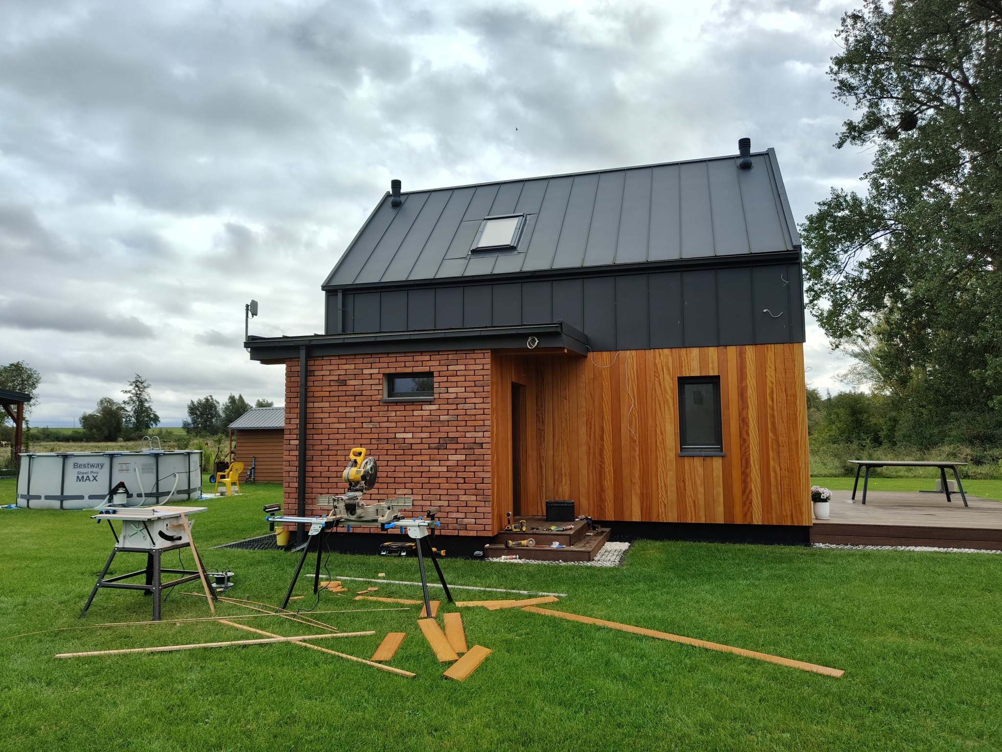Nowoczesny dom z elewacją z cegły i drewna, narzędzia do obróbki drewna na trawniku, basen w tle. Widok z zewnątrz, pochmurne niebo.