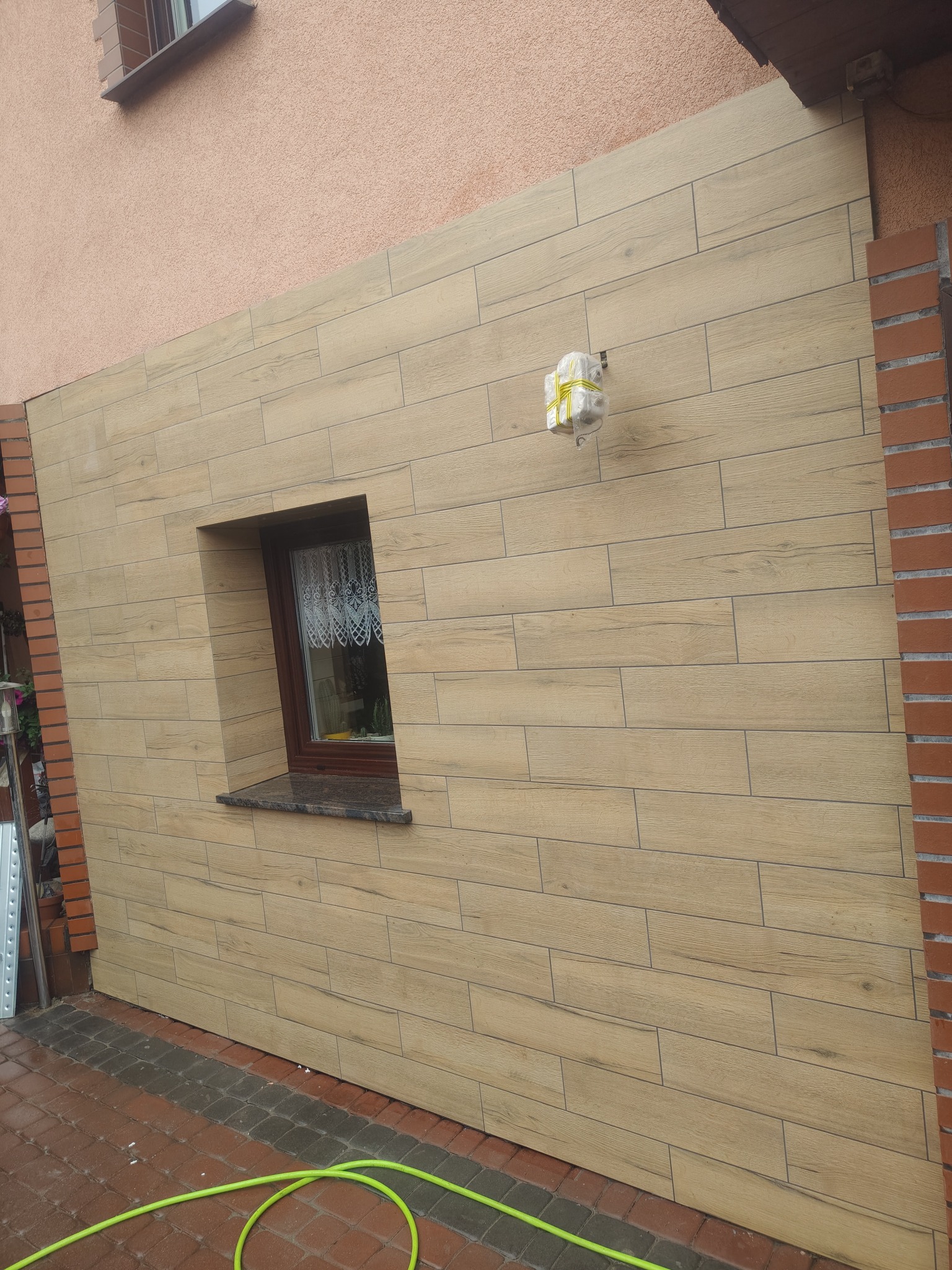 Elewacja budynku wykończona płytkami imitującymi drewno, z oknem i wystającym elementem instalacji. Widoczny fragment chodnika z kostki brukowej.