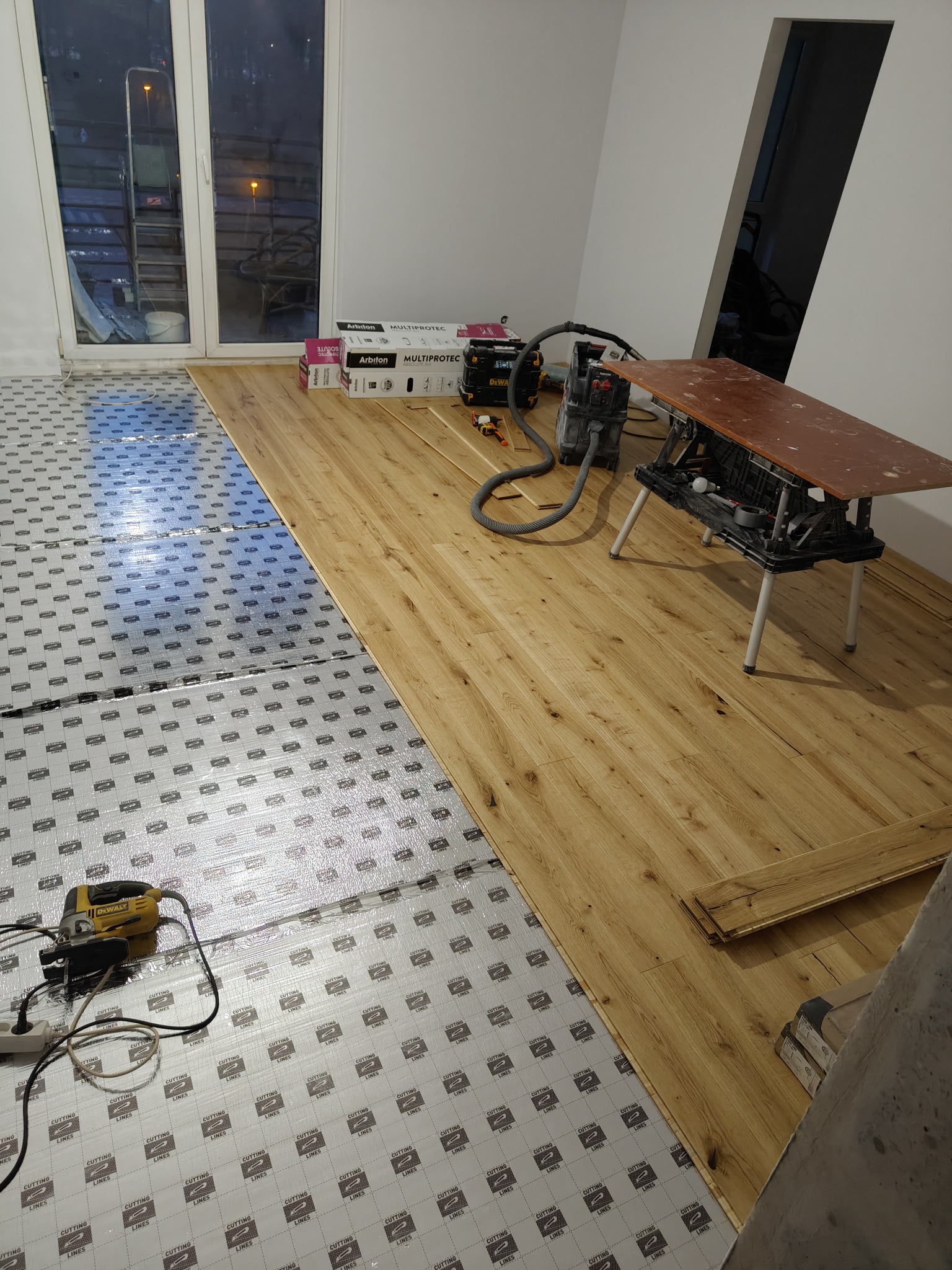 Układanie paneli podłogowych: widok na częściowo ułożone panele w odcieniach dębu, podkład izolacyjny, narzędzia (wyrzynarka, odkurzacz) i paczki paneli Arbiton.