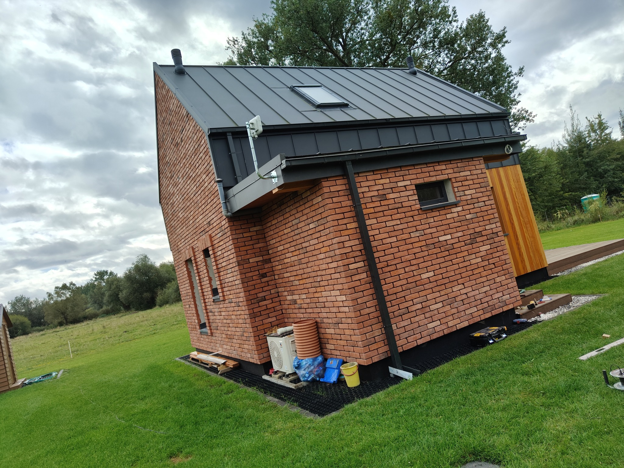 Nowoczesny dom z cegły klinkierowej i grafitowym dachem na tle zielonego trawnika i pochmurnego nieba. Widoczne detale wykończenia i elementy instalacji.