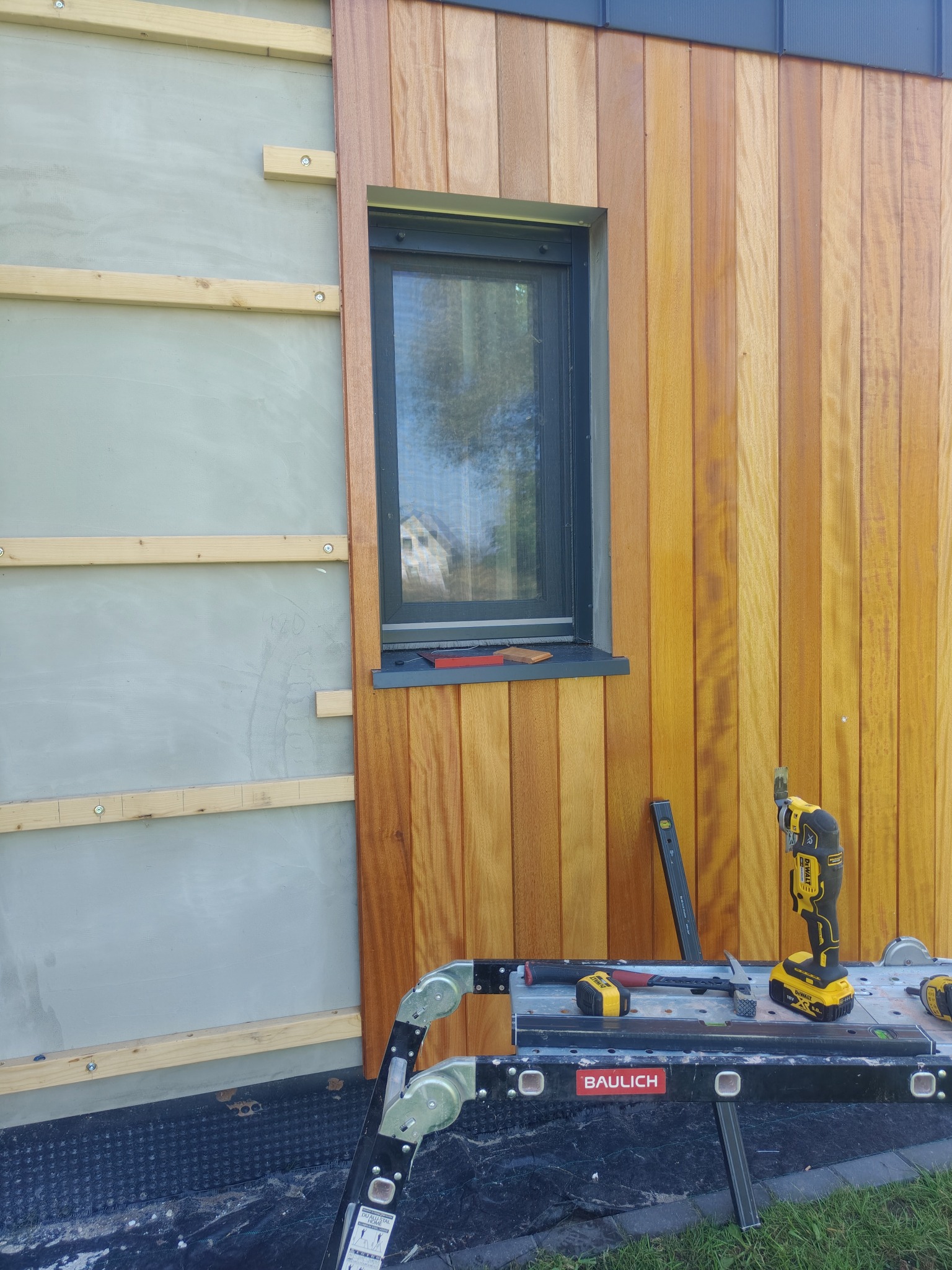 Montaż drewnianej elewacji zewnętrznej budynku. Widoczne deski, okno, rusztowanie i narzędzia. Częściowo ocieplona ściana z drewnianym rusztem.