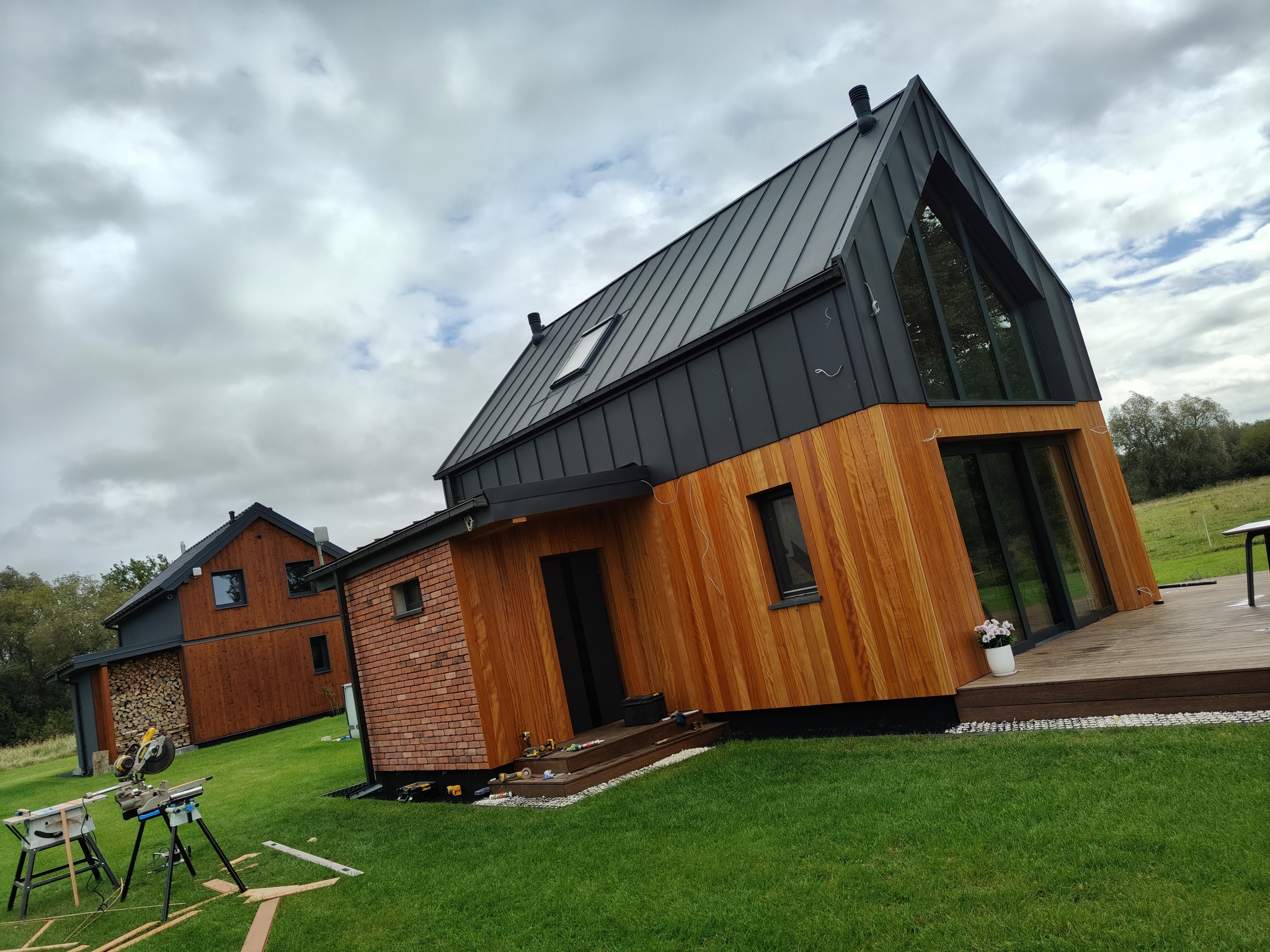 Nowoczesny dom z drewnianą elewacją i grafitowym dachem na tle pochmurnego nieba. Na pierwszym planie narzędzia budowlane na trawie, sugerujące trwające prace wykończeniowe.