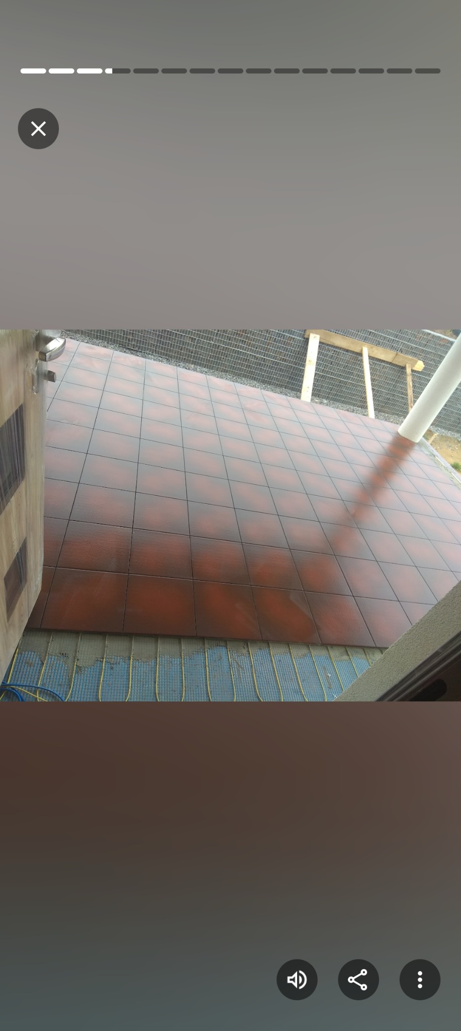 Brązowe płytki podłogowe ułożone na ogrzewaniu podłogowym, widoczne przez otwarte drzwi. Siatka i elementy konstrukcyjne w tle.