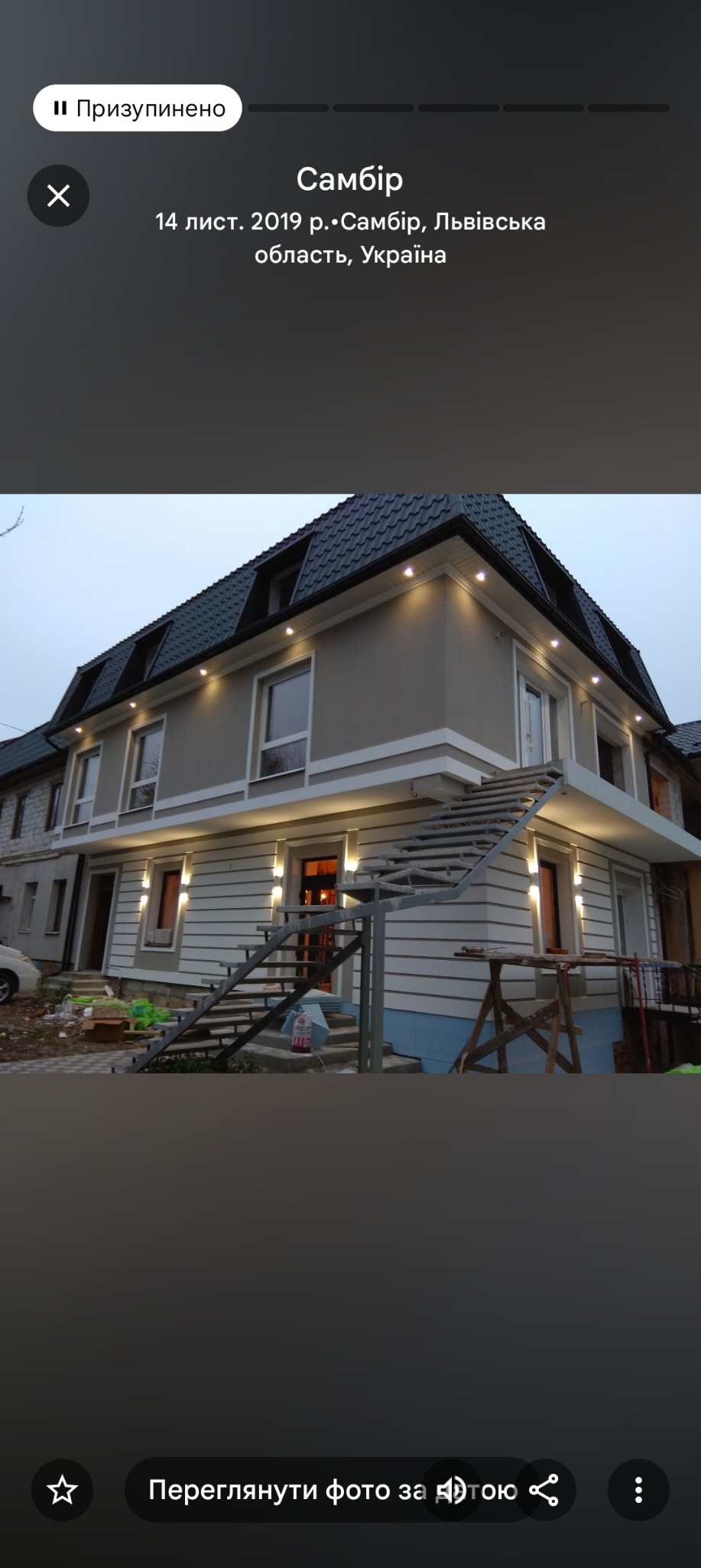 Dwupiętrowy dom z szarą elewacją i czarnym dachem, oświetlony ciepłym światłem. Metalowe schody prowadzą do wejścia. Widoczne materiały budowlane.