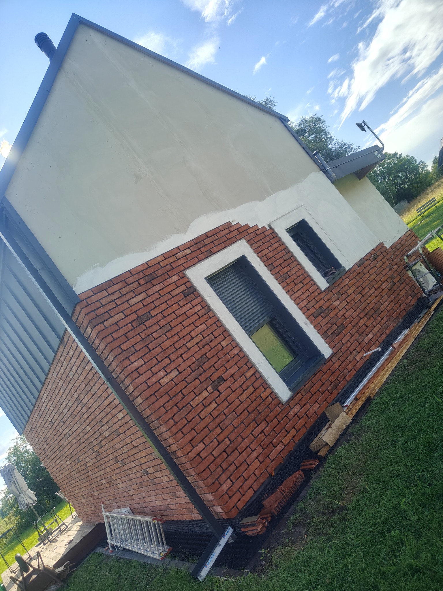 Częściowo wykończona elewacja domu z cegły klinkierowej wokół okien. Widoczne rusztowanie, materiały budowlane i trawnik. Ujęcie z dołu, lekko obrócone.