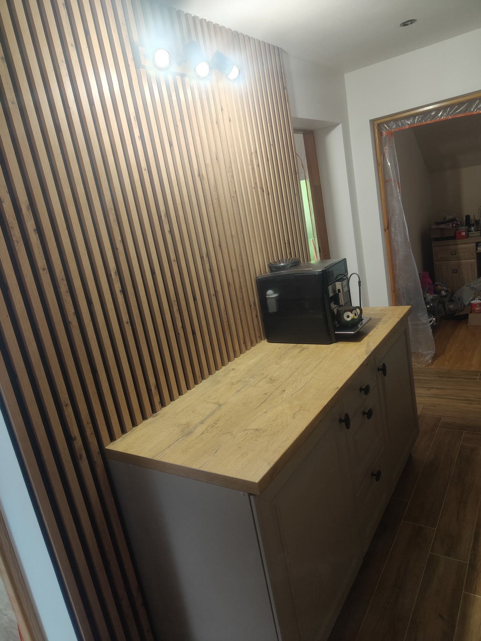 Nowoczesna aranżacja wnętrza z drewnianą ścianą z lameli i szafką z blatem imitującym drewno. Na blacie stoi ekspres do kawy. Podłoga wyłożona płytkami imitującymi drewno.