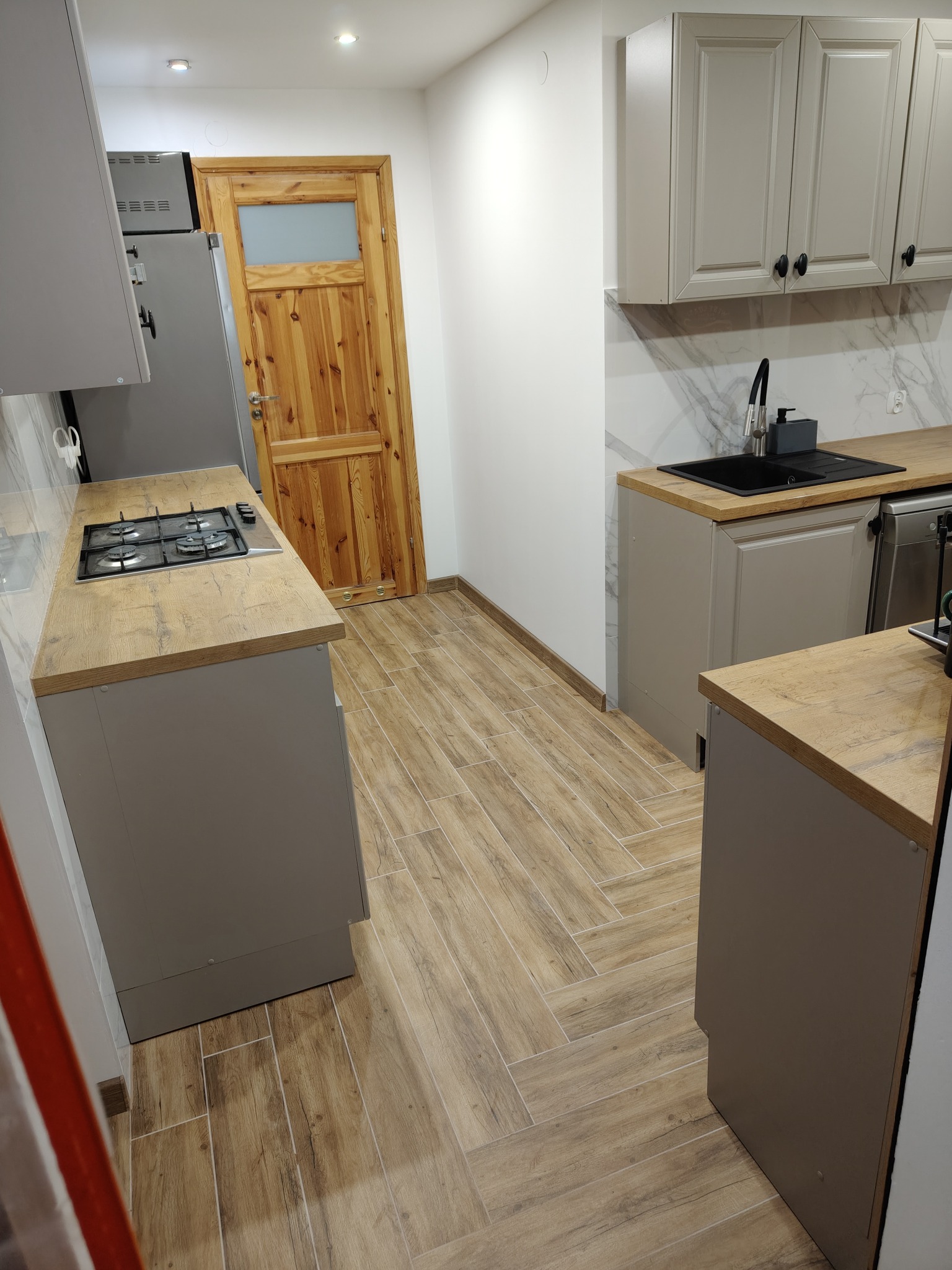 Jasna kuchnia z drewnianą podłogą ułożoną w jodełkę, szare szafki z czarnymi uchwytami i drewniany blat. Widoczny fragment lodówki i drzwi z szybą.