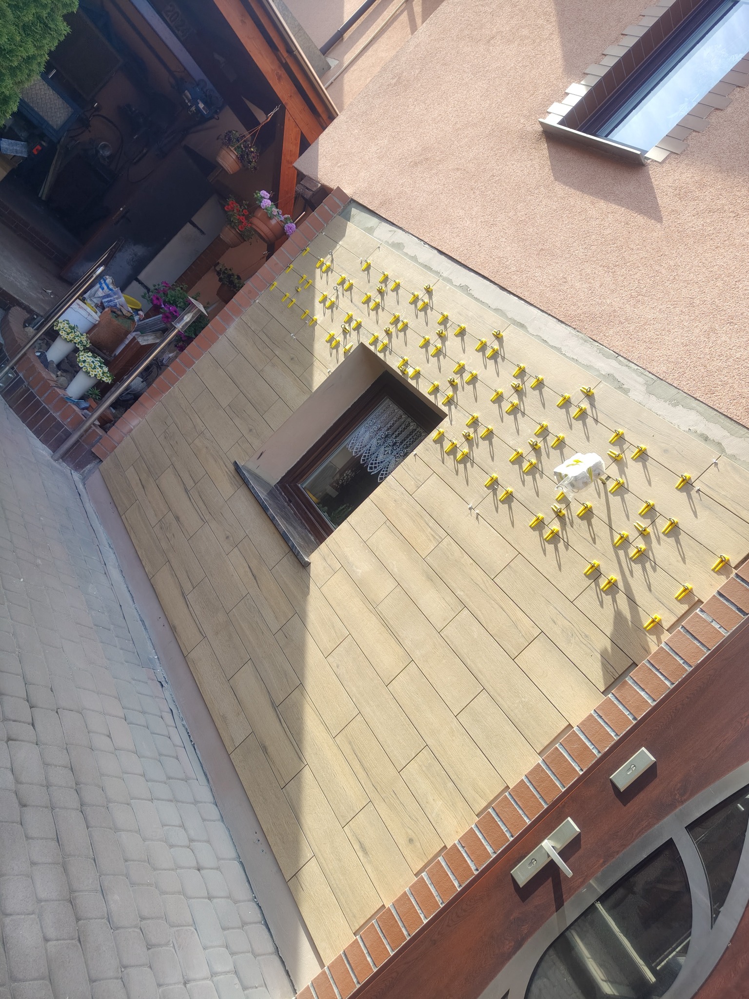 Elewacja budynku w trakcie wykańczania, z widocznymi płytkami imitującymi drewno i żółtymi klipsami poziomującymi. Okno w elewacji. Ujęcie z góry.