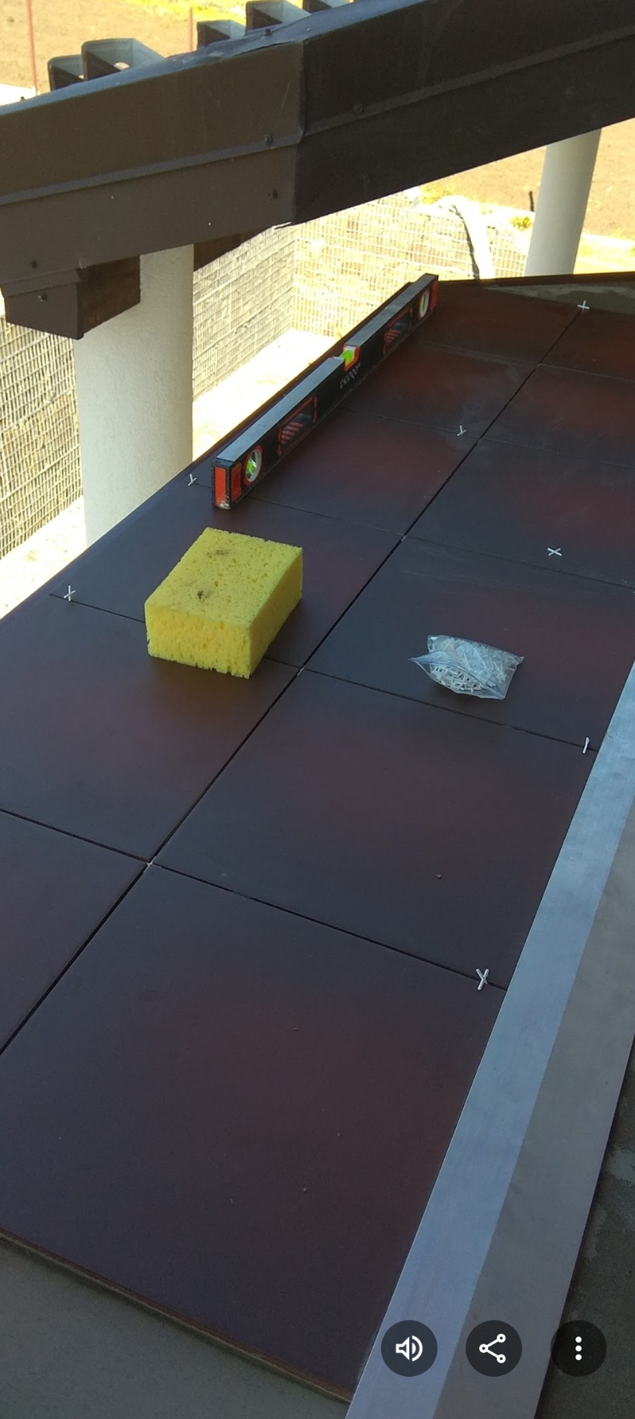 Brązowe płytki ułożone na zewnątrz, poziomica, gąbka i krzyżyki dystansowe. Widok z góry na etap prac wykończeniowych na tarasie.