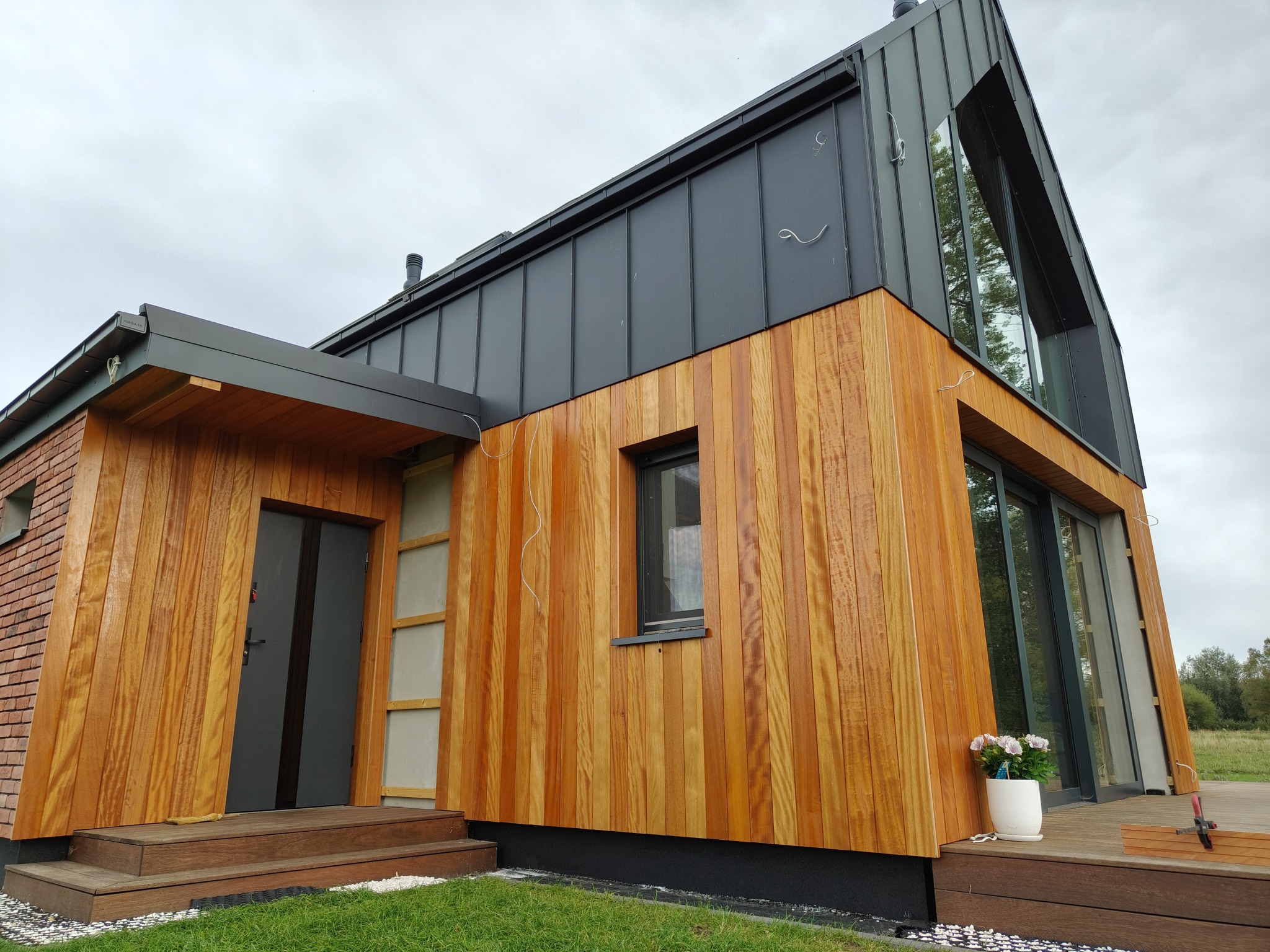 Nowoczesny dom z drewnianą elewacją i grafitowym dachem. Widoczne schody wejściowe i okno. Na tarasie donica z kwiatami. Całość w naturalnym otoczeniu.
